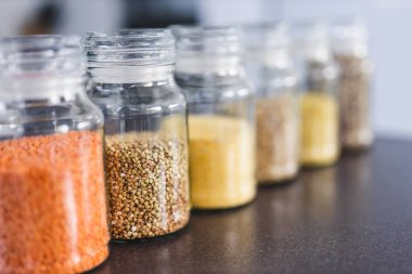 İçinde kinoa mercimek, karabuğday ve arpanın da bulunduğu farklı türde tahıl ve baklagillerin bulunduğu açık kiler kavanozları mutfak tezgahı, basit malzeme konsepti.