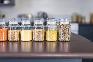 İçinde kinoa mercimek, karabuğday ve arpanın da bulunduğu farklı türde tahıl ve baklagillerin bulunduğu açık kiler kavanozları mutfak tezgahı, basit malzeme konsepti.