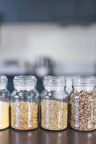 İçinde kinoa mercimek, karabuğday ve arpanın da bulunduğu farklı türde tahıl ve baklagillerin bulunduğu açık kiler kavanozları mutfak tezgahı, basit malzeme konsepti.