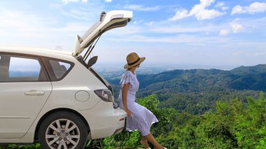 Dağlarda beyaz hatchback araba bagajında dinlenen ve mavi gökyüzü ve beyaz bulut arka planıyla izleyen bir kadın. Gezgin araba konsepti.