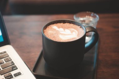 Latte art hot mocha coffee with milk latte art on top of black ceramic glass set on table in coffee shop next to computer notebook in during the day clipart