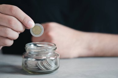 Man hand putting ten baht Thai coins into a glass bottle. Save money concept