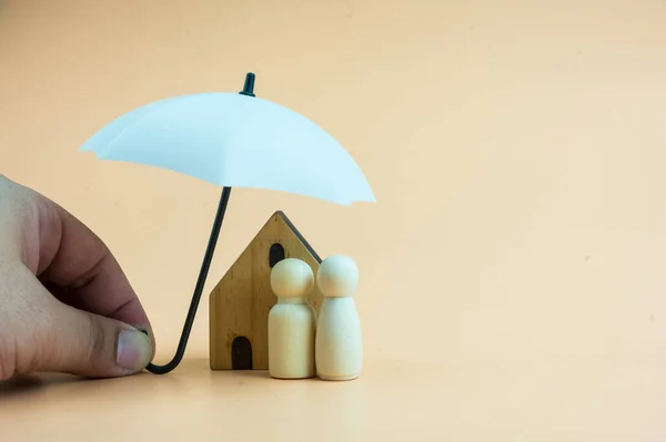 Family of wooden dolls and home are under a white umbrella, protecting wooden peg dolls and home, planning, saving families, preventing risks and crises, and insurance concepts.