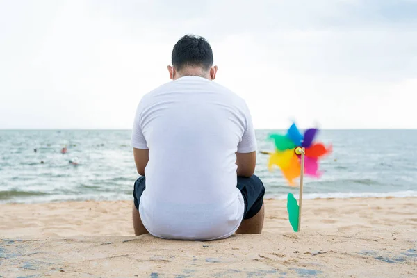 Rear view of lonely man wears a white shirt and sitting on the beach near pinwheel toy and using a phone. The ultimate place to relax and restart