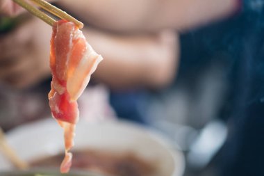 Hand uses chopsticks to pick up raw bacon to grilled. Close up