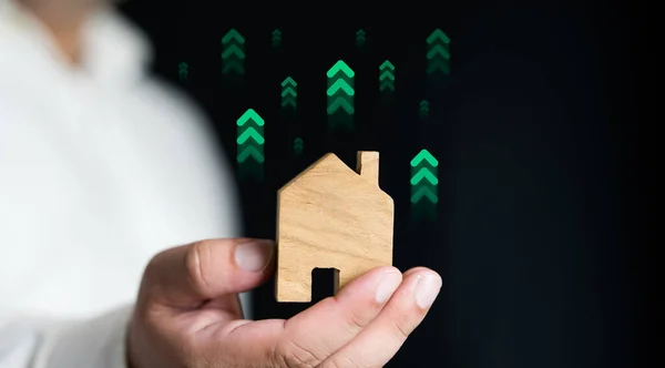 Man holding model house with green arrows up on a black background. Interest rate home up, affordable alternative price, mortgage home, home financial and investment concept.