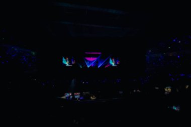Stadyumda bulanık kalabalık kutlama yapıyor. Parlak sahne ışıkları, kalabalık dans pisti. Konserde telefon ışıkları. Bando mavisi siluet kalabalık.