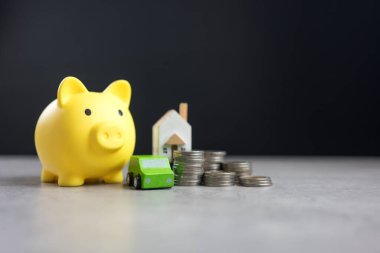 Mülk konsepti, sigorta, yatırım, mortgage, kar, mülk, kira, finans ve tasarruf. Domuz kumbarası, ahşap bir evin, yeşil bir arabanın ve bozuk para stoğunun yanında sarıdır..