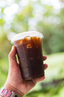 Buzlu americano plastik cam ellerde bahçe arka planı ve bokeh ışığı var.