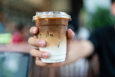 Adamın elinde plastik bardak buzlu kahve var. Üst katmanda esspeso var. Altında taze süt, seçici odak noktası..