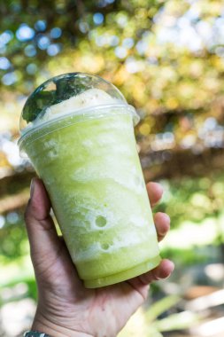 Yeşil sebze ve meyve suyu dolu şeffaf plastik bardak ve arka planda bulanık bir kapak.