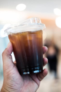 Şeffaf plastik kapaklı bir kupa ve elinde buzlu bir Americano.
