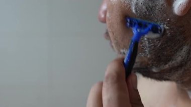 Yakışıklı Asyalı bir erkeğin banyoda traş bıçağı ile sakalını tıraş edişini.