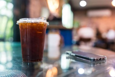 Buzlu siyah kahveyle dolu plastik bir fincan parlak ve cep telefonunun cam masasında duruyor. Arka planı bulanık modern bir kafe..