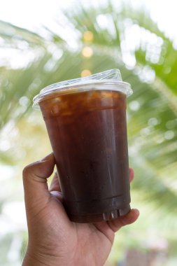 Doğanın bulanık arka planında plastik bir bardak Ameracano kahvesi tutuyor.