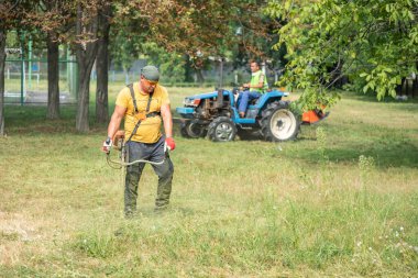 Erkek bahçıvan elektrikli çim biçme makinesi ya da petrol biçme makinesiyle çim biçiyor erkek bahçıvan da bir çim biçme makinesi ile arka planda çim biçiyor. Bölgenin bakımı, çim biçme. Bahçe bakımı aletleri ve ekipmanları.