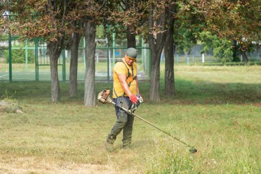 Adam benzin tırpanıyla çim biçiyor. Erkek bahçıvan parkta elektrikli çim biçme makinesi ya da benzin biçimiyle çim biçiyor. Bahçe bakımı aletleri ve ekipmanları.