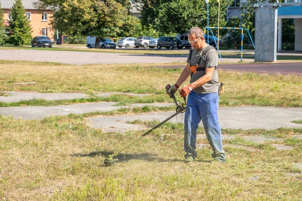 İşçi benzin tırpanıyla çim biçiyor. Bahçıvanlık malzemeleri ve ekipmanlar. Bahçıvan, şehir caddesi boyunca elektrikli çim biçme makinesiyle çim biçiyor. Çim biçme makinesiyle çim biçme işlemi.