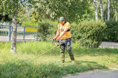 Güneşli bir günde dışarıda çim biçme makinesiyle çim biçen genç bir işçi. Bir erkek bahçıvan, halka açık bir parkta ya da cadde boyunca çim biçme makinesiyle çimenleri biçer..