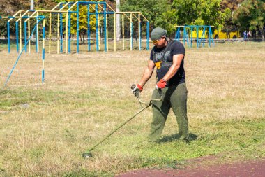 Erkek bahçıvan, spor sahasında çim biçme makinesi ve elektrikli çim biçme makinesi ile çimen biçiyor. İşçi otları benzin tırpanı ile biçiyor. Bahçe bakımı aletleri ve ekipmanları.