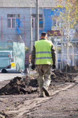 İş kıyafetleri içinde bahçıvan şehir parkında ya da meydanda yeni dikilmiş bir ağaç sokağının yanında yürüyor. Şehir sokaklarının peyzajı, güneşli bir bahar gününde ağaçların filizlendiği bahçe..