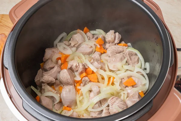 Frying meat with onions and carrots in a multicooker pan. Modern, bright multi cooker with products. Cooking pilaf with meat in multicooker. Close-up top view of multi cooker with vegetables and meat.