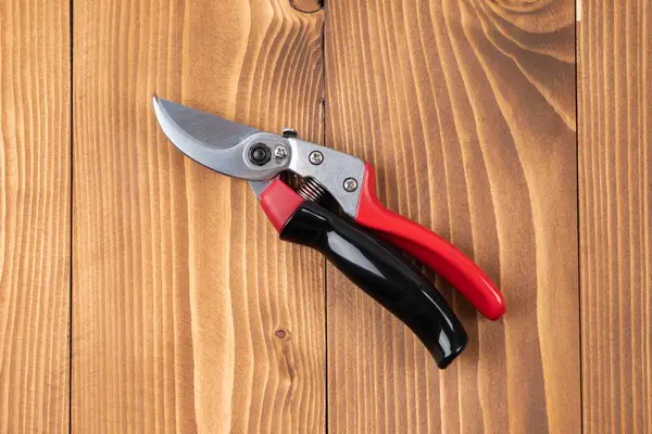Steel gardening secateurs, scissors tool with red and black grip for pruned of plants and flowers garden work, on a wooden table background. Close state. Top view. Close-up.