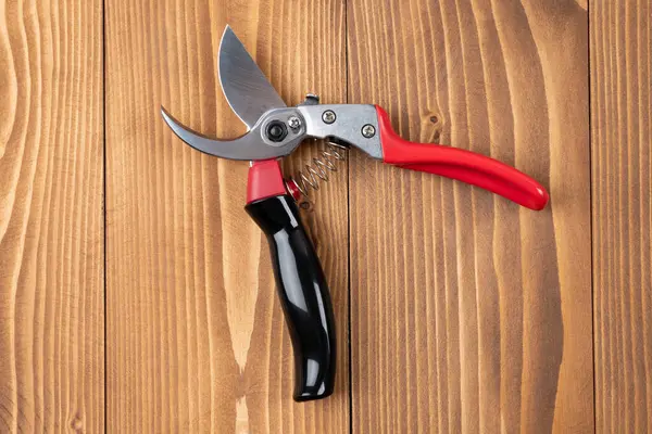 Steel gardening secateurs, scissors tool with red and black grip for pruned of plants and flowers garden work, on a wooden table background. Open state. Top view. Close-up.
