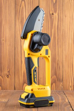 Small handheld lithium battery powered chainsaw for trimming, cutting trees or bushes branches, on a wooden table background. Vertical. Close-up. clipart