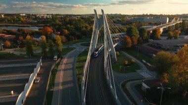 Polonya, Krakow 'daki tramvay hareketiyle birlikte asma köprü manzarası için insansız hava aracı gönderin. Köprü yapısı ve toplu taşımacılık günbatımında hareket halinde, boş asfalt yol ve aşağı park