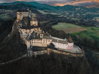 Slovakya 'da ormanlı yüksek kayalıklardaki ortaçağ Orava Kalesi' nin hava panoramik görüntüsü. Romanesk ve Gotik taş kaleli karanlık akşam manzarası dramatik gökyüzü, dağlar ve yeşil tarlalar altında