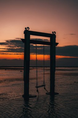 Sahilde gün batımında bir salıncağın silueti. Titreşen gökyüzü sakin suya yansıyarak huzur veren sakin ve resmedilmeye değer bir sahne yaratır..