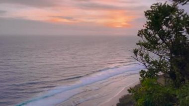 Gün batımında okyanus ufkunda, ön planda hafif dalgalar ve yemyeşil yeşillikler var. Kıyı günbatımının huzur veren güzelliğini vurgular.