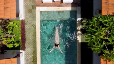 Yüzme havuzunun en üst manzarası. Etrafı yemyeşil bitkilerle ve modern mobilyalarla çevrili bir kadın yüzüyor. Sahne bir rahatlama ve lüks anı yakalar..
