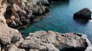 Bir insan berrak ve sakin bir okyanusun kenarında bir kayanın üzerinde duruyor. Görüntü, kıyı manzarasının sakin güzelliğini, köpüklü su ve engebeli kaya oluşumlarıyla yakalıyor. Yalnızlık ve sükuneti vurguluyor..