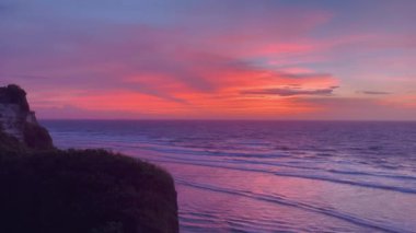 Sahildeki kayalıklar ve okyanus dalgaları üzerinde canlı renklerle dramatik günbatımı manzarası. Alacakaranlıkta doğanın nefes kesici güzelliğini vurgular.