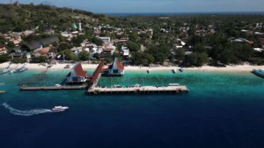 Bali 'deki canlı bir sahil kasabasının temiz mavi sulara uzanan rıhtımı görüntüsü. Hareketli bölgede renkli binalar, tekneler ve canlının tadını çıkaran kumsala gidenler var.