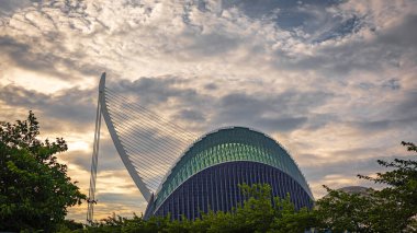 Modern bir mimari mucize, gün batımında bulutları olan dramatik bir gökyüzüne karşı duruyor. İnşa çizgileri ve yenilikçi tasarım altın ışıkla vurgulanır..