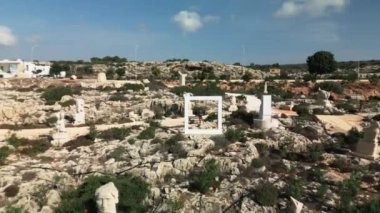 Kamera geriye doğru hareket ederken çerçevenin ortasında duran bir adam Kıbrıs Rum Kesimi 'nin Ayia Napa kentindeki geniş heykeller parkının manzaralı ve kayalık bir manzaraya sahip çeşitli heykellerini gözler önüne seriyor.