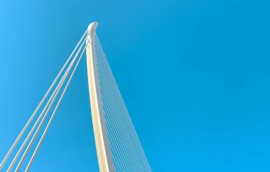 Açık mavi gökyüzüne karşı modern bir asma köprünün alçak açılı görüntüsü, yapısal mühendisliği, parlak tasarımı ve kentsel mimariyi vurguluyor..