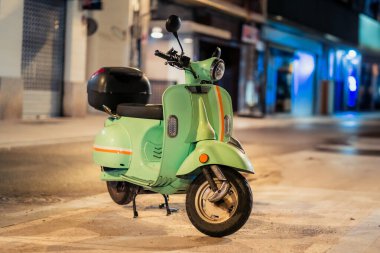 Büyüleyici yeşil bir scooter, hareketli bir şehir caddesinde, gece lambalarıyla güzelce aydınlatılmış, gelişigüzel park edilmiş.