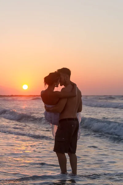 해변에서 부부가 늦여름의 황혼무렵 드레스와 반바지를 맨발로 놀리고 키스를 로열티 프리 스톡 이미지