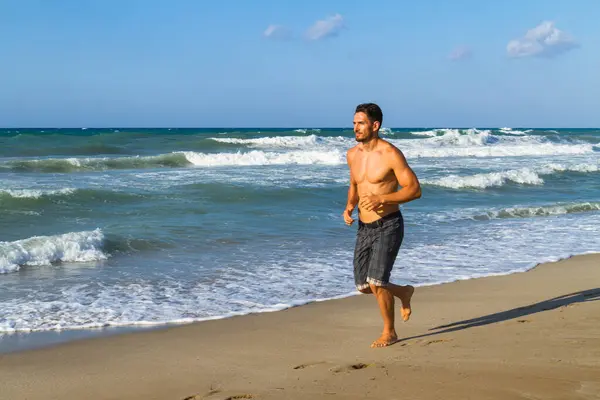 stock image Young man in his twenties jogging along the shoreline on a sandy beach, late afternoon in the summer.