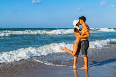 Bikinili ve şortlu mutlu genç çift yazın tadını çıkarıyor alacakaranlıkta kumsalda, temiz hava soluyor, öpüşüyor ve birbirlerine sataşıyorlar.