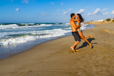 Bikinili ve şortlu mutlu genç çift yazın tadını çıkarıyor alacakaranlıkta kumsalda, temiz hava soluyor, öpüşüyor ve birbirlerine sataşıyorlar.