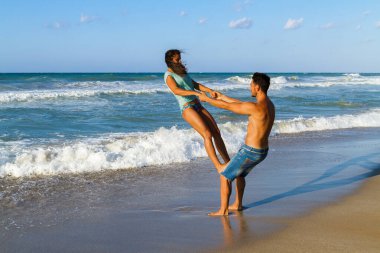 Bikinili ve şortlu mutlu genç çift yazın tadını çıkarıyor alacakaranlıkta kumsalda, temiz hava soluyor, eğleniyor ve birbirlerine sataşıyorlar.