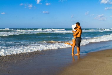 Bikinili ve şortlu mutlu genç çift yazın tadını çıkarıyor alacakaranlıkta kumsalda, temiz hava soluyor, öpüşüyor ve birbirlerine sataşıyorlar.