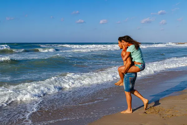 Bikinili ve şortlu mutlu genç çift yazın tadını çıkarıyor alacakaranlıkta kumsalda, temiz hava soluyor, eğleniyor ve birbirlerine sataşıyorlar.