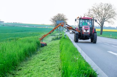 11 Mayıs 2022 Crudim, Çek Cumhuriyeti. Döner çim biçme makinesi olan bir traktör yol kenarında yeşil çim biçer. Kaldırımları temizlemek, yol kenarlarını biçmek....