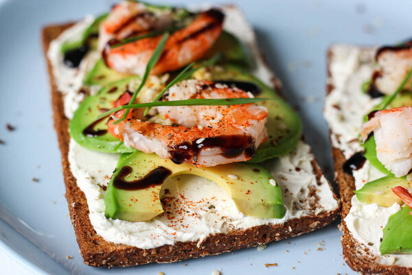 Вкусный сэндвич с открытым лицом со сливочным сыром, авокадо, креветками и бальзамической глазурью на цельнозерновом хлебе. Он увенчан кунжутом, копченой паприкой и пшеничной микрозеленью. Идеальная закуска из морепродуктов или здоровая идея позднего завтрака